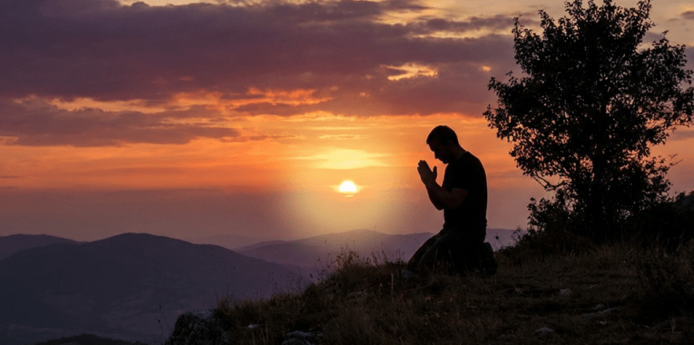 Silhouette of a man kneeling in prayer on a hilltop during a vibrant sunset.