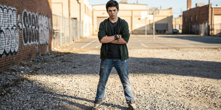 Teenager with crossed arms standing on gravel near a basketball court with graffiti-covered wall