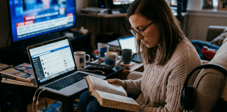 Woman sitting on couch reading book with laptop, phone, televisions, and magazines around