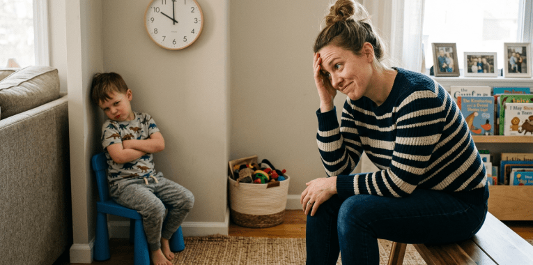 Toddler sitting with arms crossed in timeout while parent watches with hand on forehead