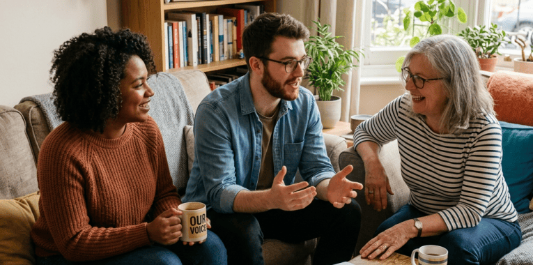 Three adults discuss community matters beside a “Community Event” sign and “OUR VOICE” mug.